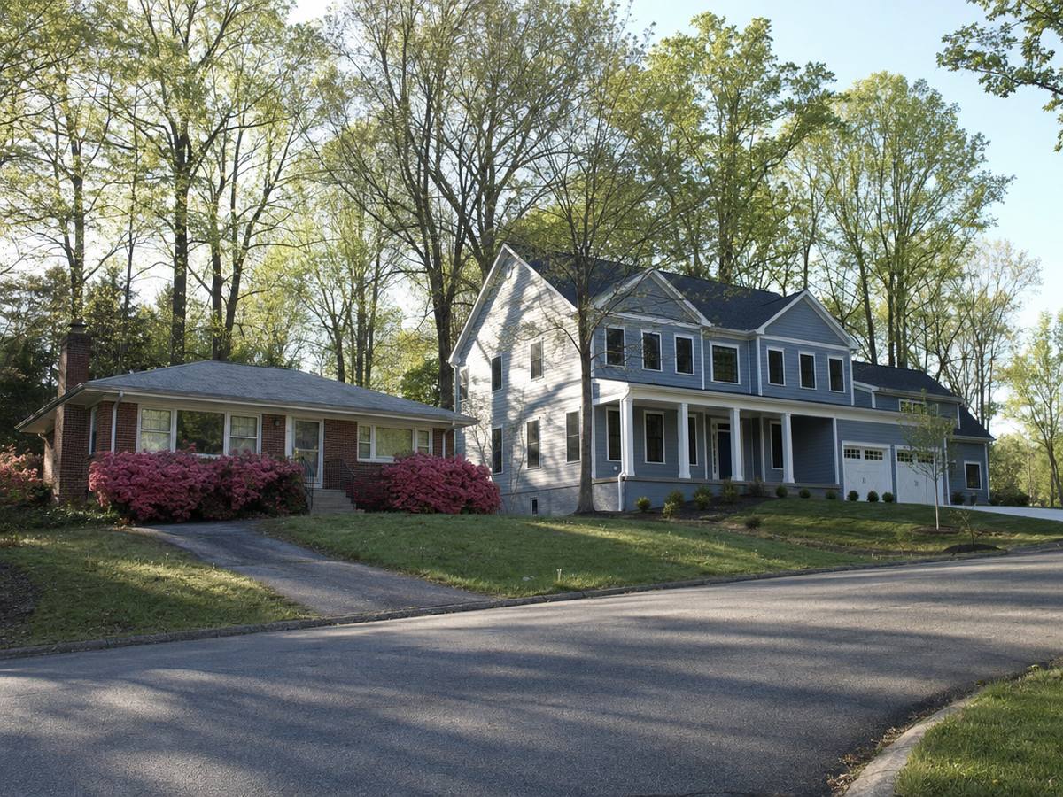 A small red-brick rambler with pink azaleas beside a large new blue-gray colonial with a front porch on a wooded, sloping street.