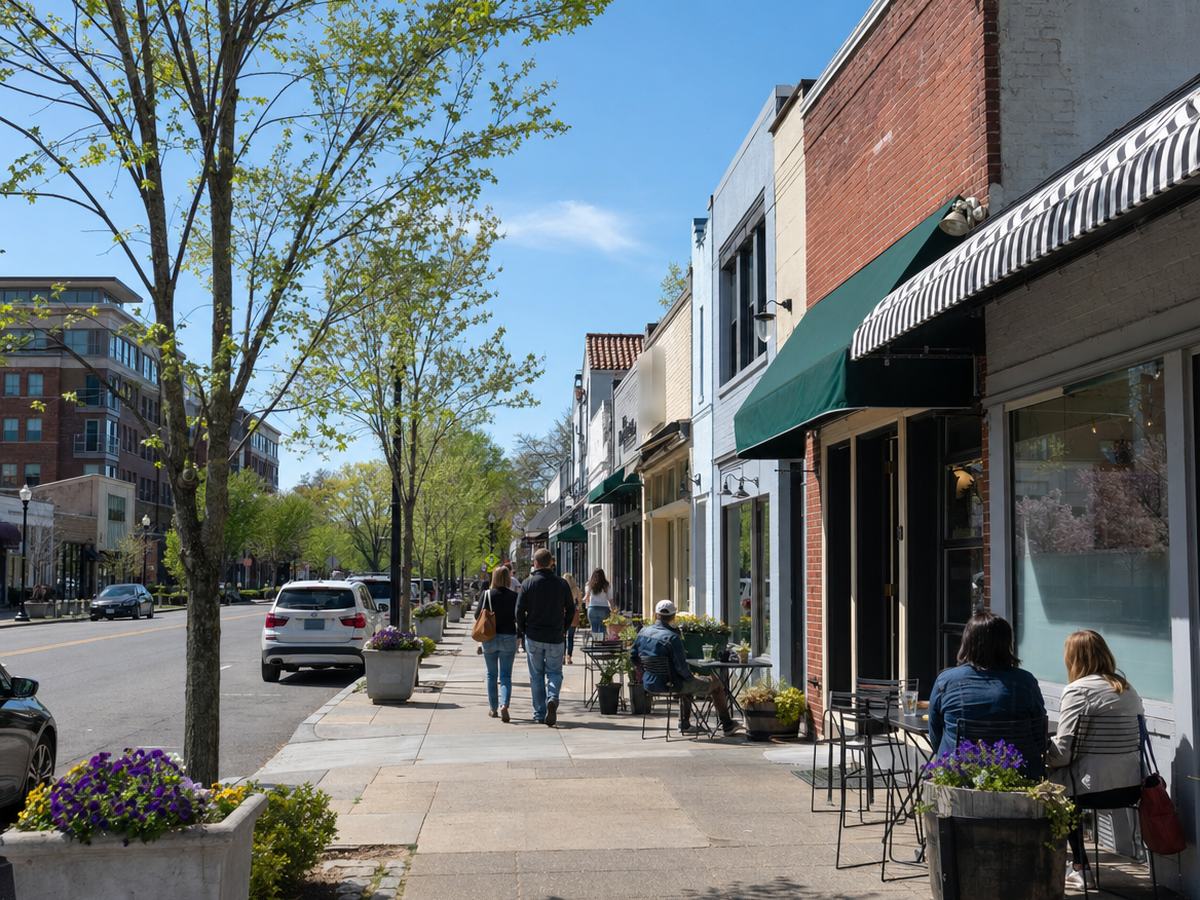 Mount Vernon Avenue in Del Ray with low brick and painted storefronts, awnings, sidewalk cafe tables, planters and young street trees, with a few people walking.